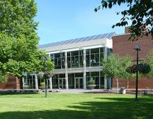 Brick building with glass entrance and solar panels on roof. Green grass and trees in front.
